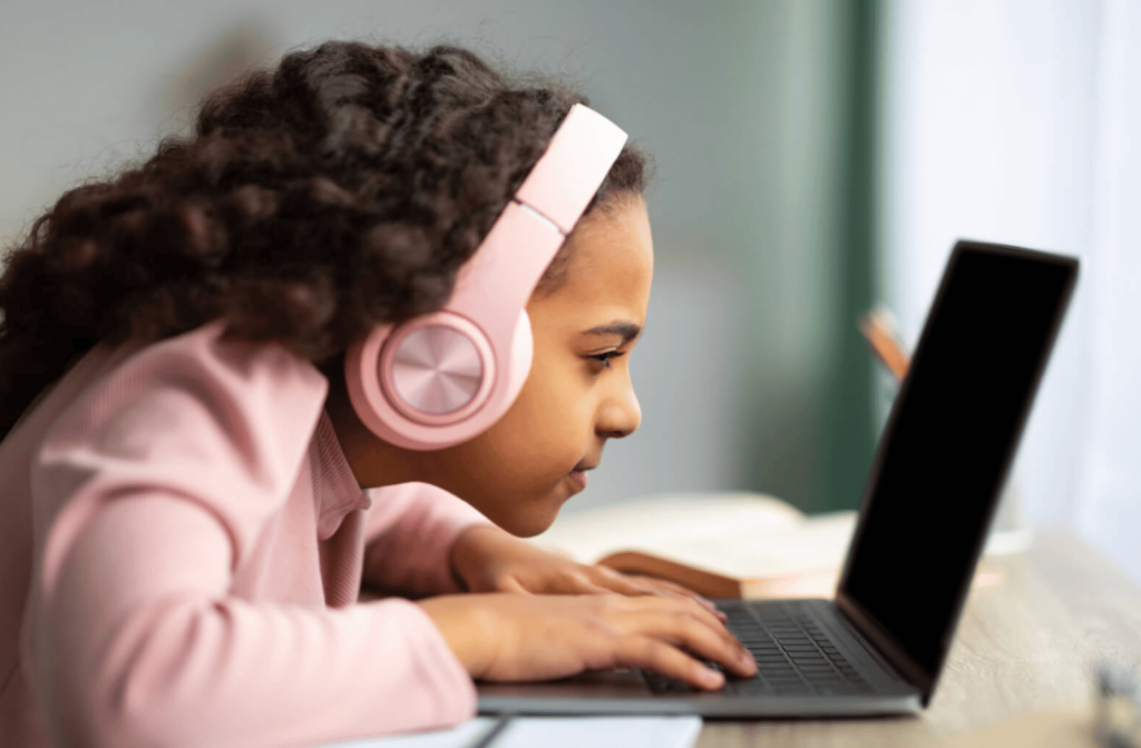 A young schoolgirl wearing pink headphones is sitting at a table too close to her laptop screen.
