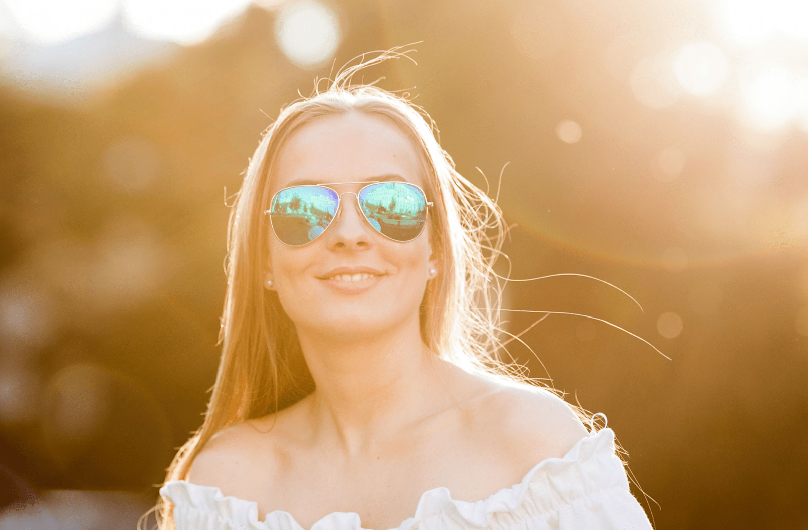 A young woman smiling while standing outside and wearing polarized sunglasses to protect her eyes.