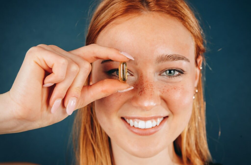 A woman holding a Omega-3 supplement close to her right eye.
