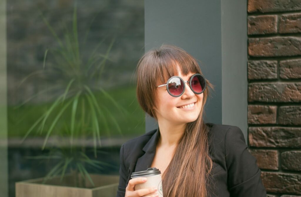 A young adult relaxes outside with a coffee while a trendy pair of sunglasses covers their eyes.