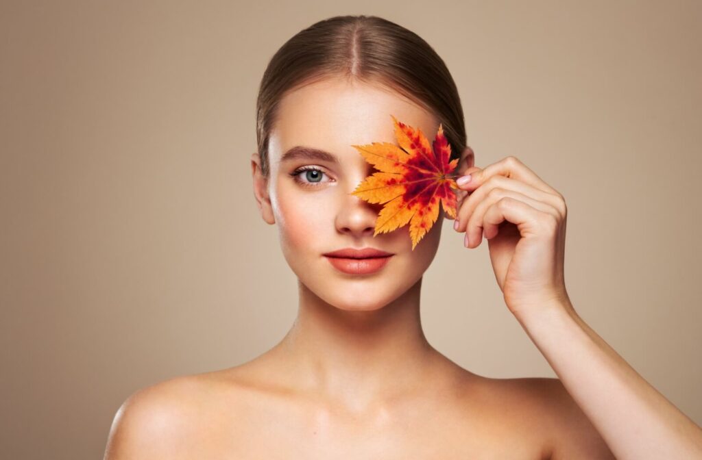  Woman holding a colorful autumn leaf over one eye, with smooth skin and natural makeup.