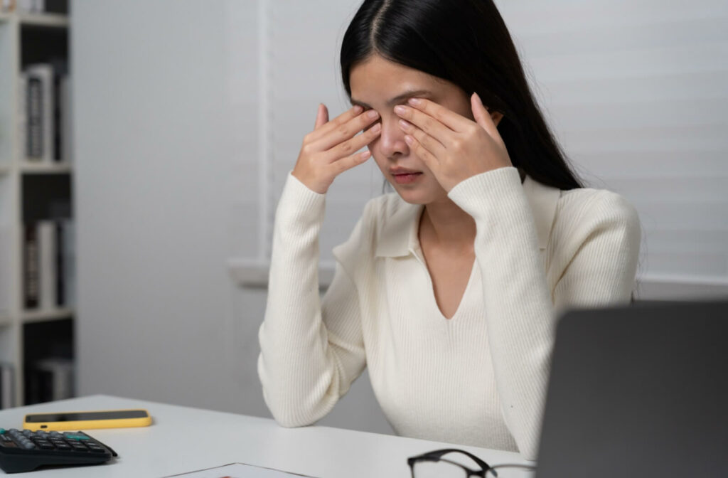 A person sitting at a desk rubs her tired eyes with both hands, showing signs of eye strain or dryness.